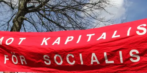 Röd banderoll med vit text: "MOT KAPITALIS FÖR SOCIALIS". En demonstration som visar vad socialism betyder.