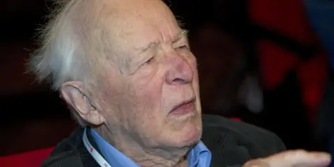 Äldre man med vitt hår, klädd i blå skjorta och mörk kavaj. Han bär ett band runt halsen med texten "Vänsterpartiet".