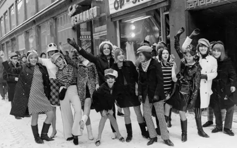 Ungdomar i trendiga kläder utanför en op-shop i Sverige 1960. Snö på marken och glada miner.