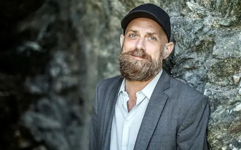 Man med skägg och keps, i grå kavaj, står mot en stenvägg. Kanske en del av en förintelsen serie på SVT.