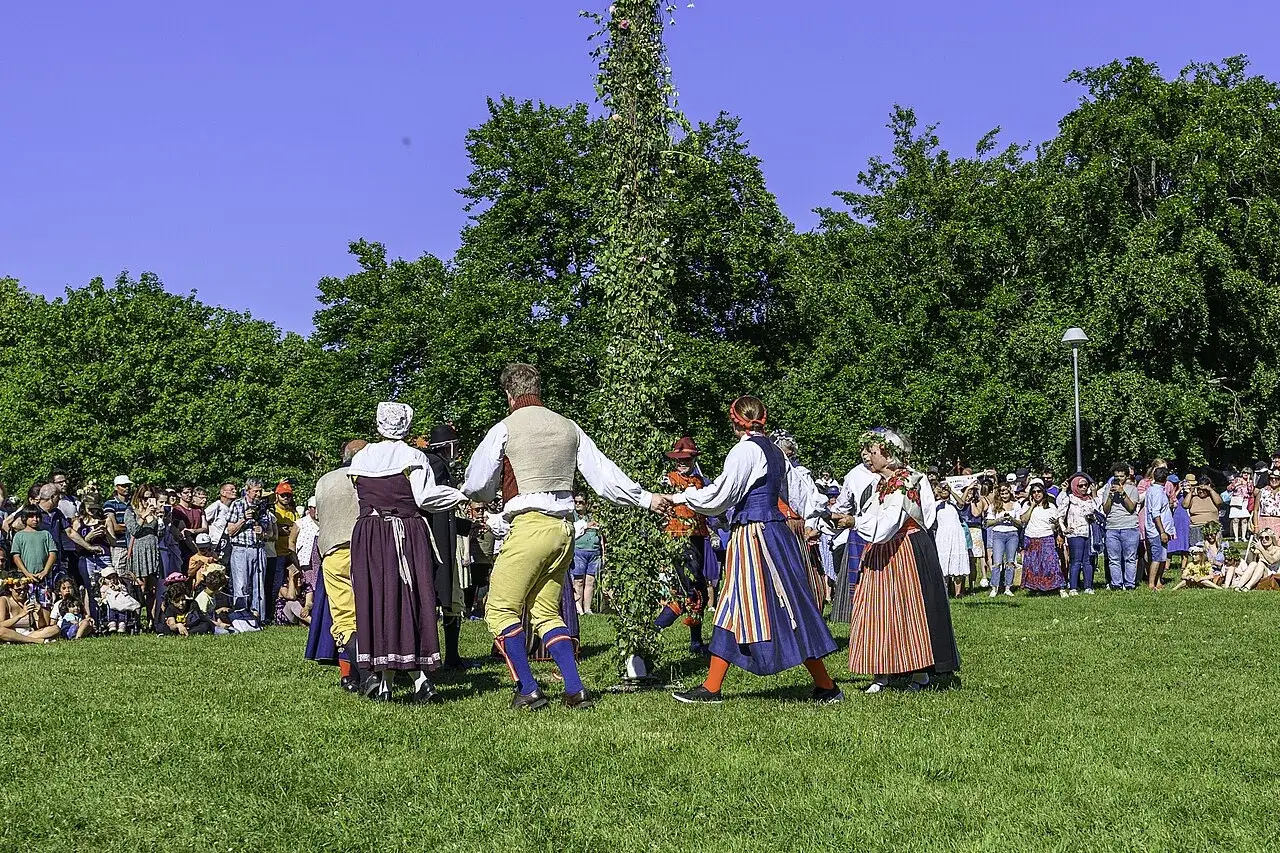 Midsommarhistoria: M&auml;nniskor i folkdr&auml;kter dansar runt en midsommarst&aring;ng p&aring; en gr&ouml;n gr&auml;smatta, omgivna av en publik.