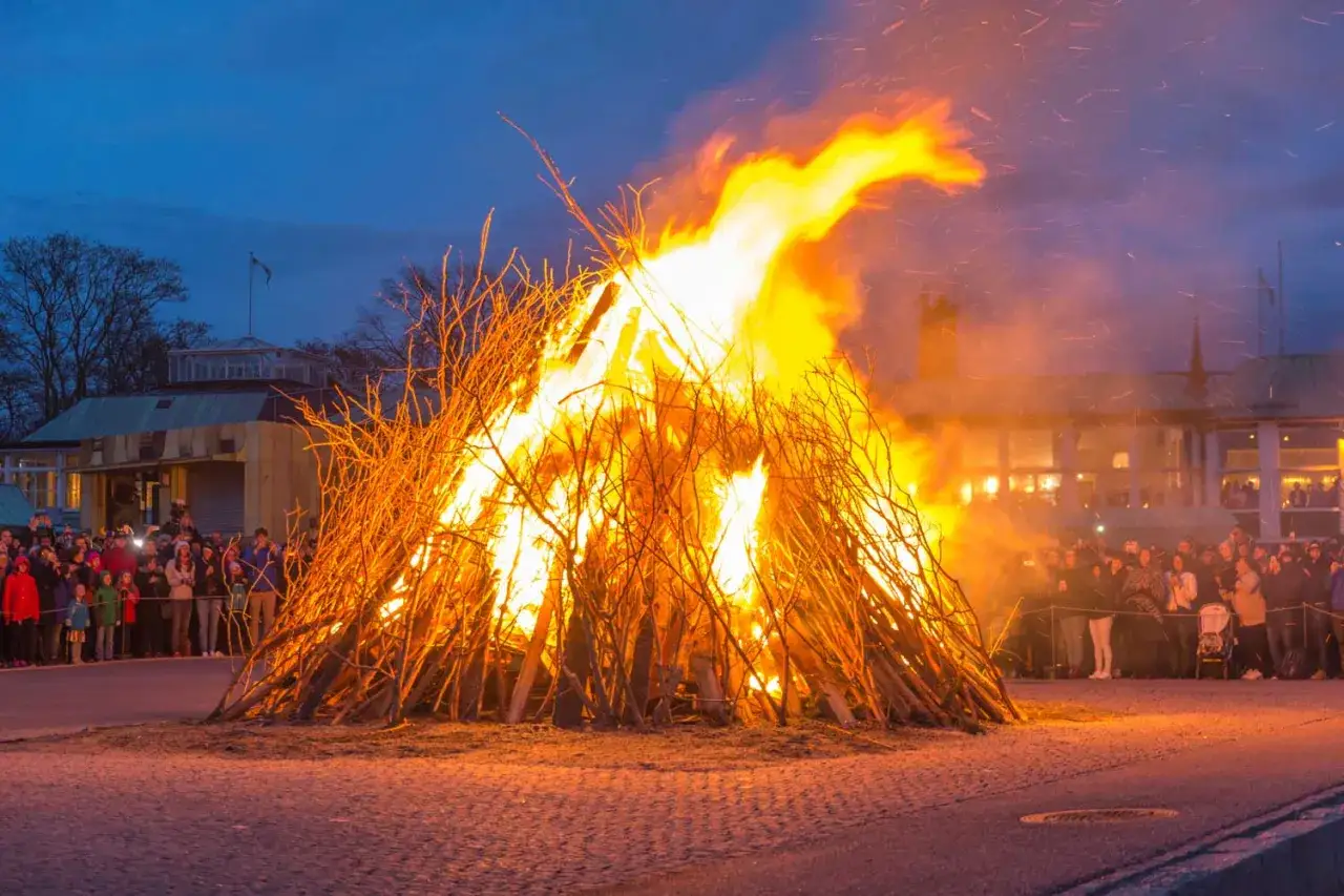 Ett stort b&aring;l flammar upp under ett valborgsfirande. M&auml;nniskor samlas f&ouml;r att se p&aring; elden och fira v&aring;rens ankomst.