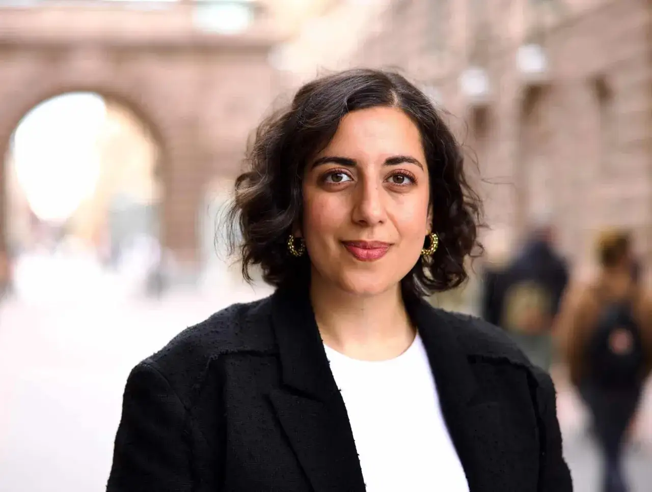 En kvinna med mörkt lockigt hår och stora örhängen, klädd i en svart kavaj, poserar utomhus. Hon ser ut som en framstående person, kanske folkpartiet partiledare.