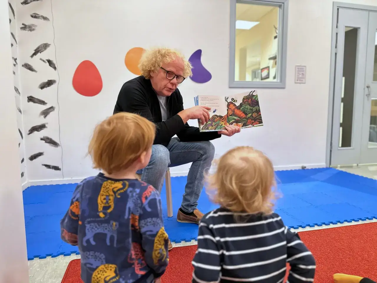En man med lockigt h&aring;r l&auml;ser en bok f&ouml;r tv&aring; sm&aring; barn. Bilden visar en humlan djojj p&aring; en morot i boken.