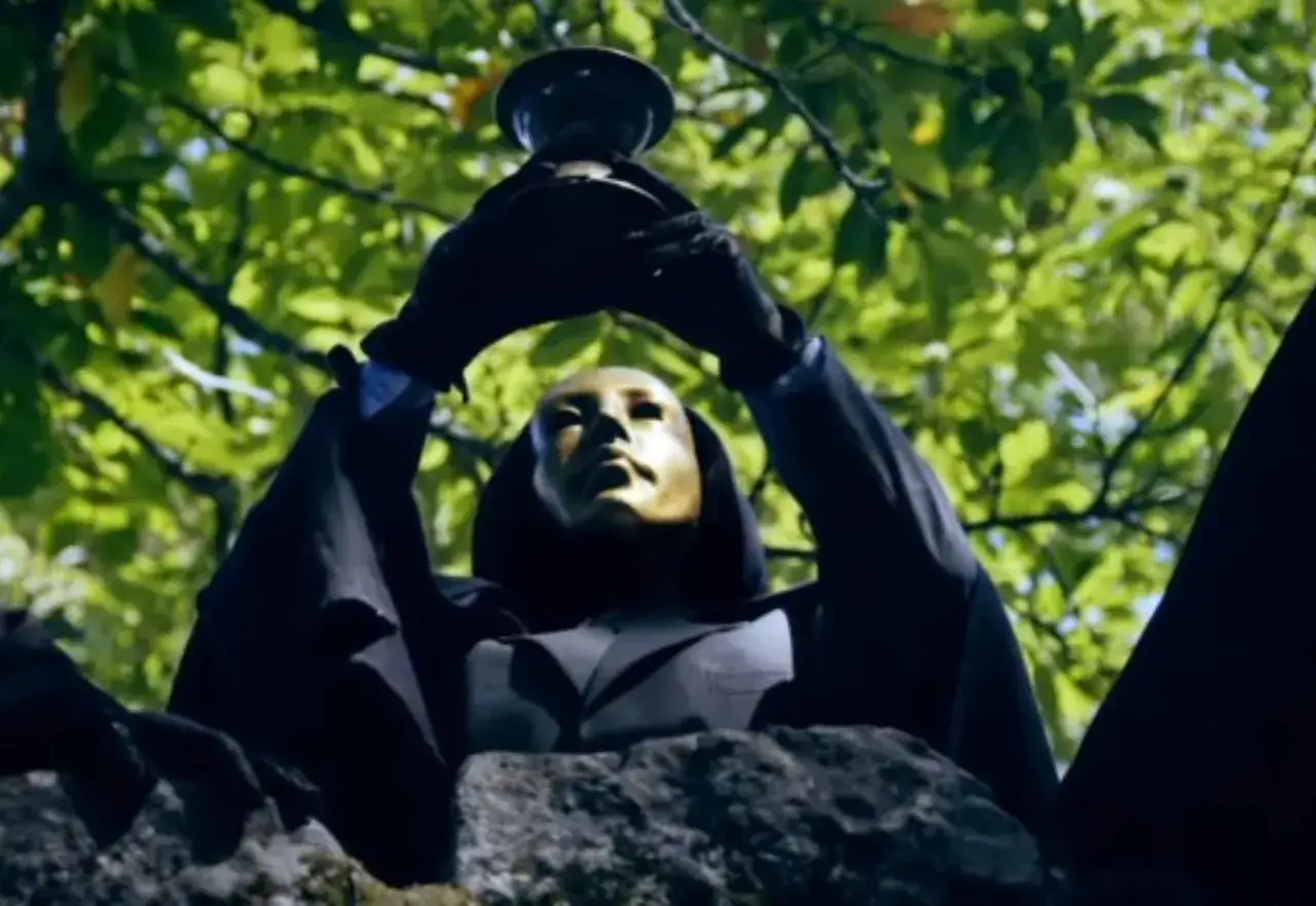 En figur med gyllene mask och svart mantel h&aring;ller upp en b&auml;gare, som en hemlighetsfull scen fr&aring;n 