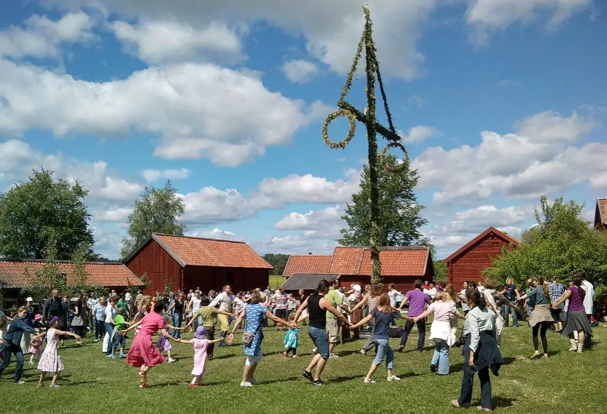 Midsommarst&aring;ngens betydelse firas med ringdans runt den l&ouml;vade st&aring;ngen. M&aring;nga m&auml;nniskor samlas p&aring; en gr&ouml;nskande &auml;ng, omgivna av r&ouml;da hus under en bl&aring; himmel med vita moln.